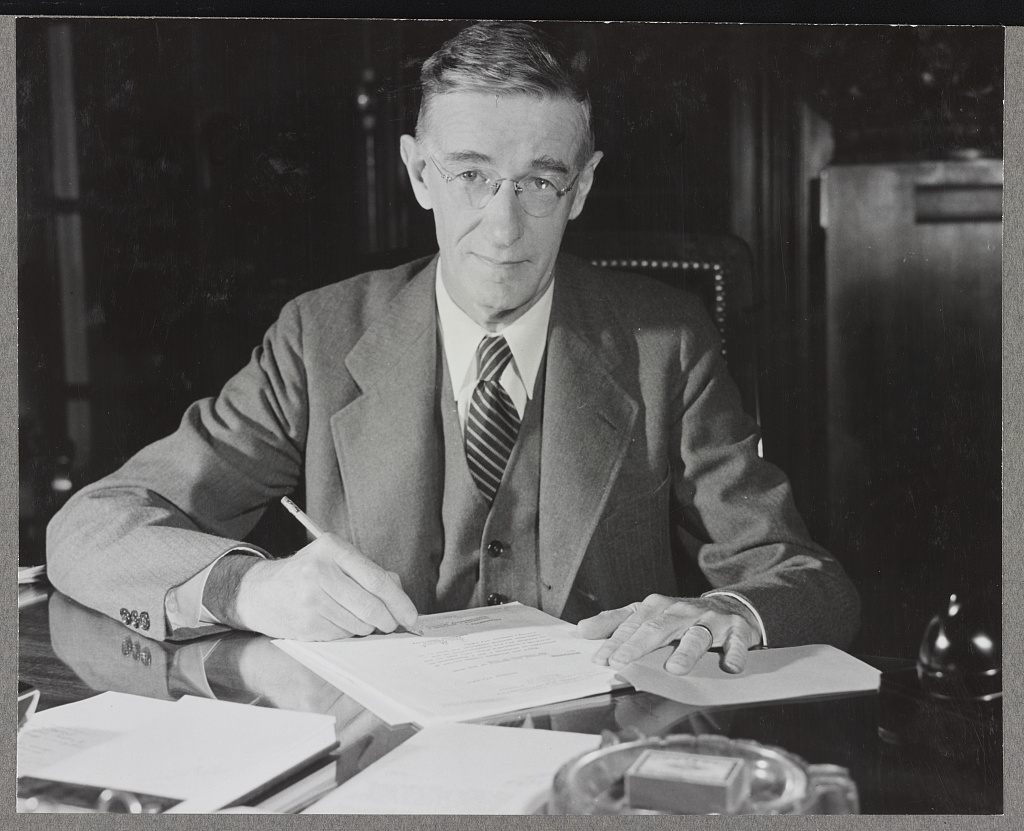 Vannevar Bush, in a three-piece suit and round glasses, sits at a polished desk piled with papers, pen in hand, looking into the camera. Black and white photograph.