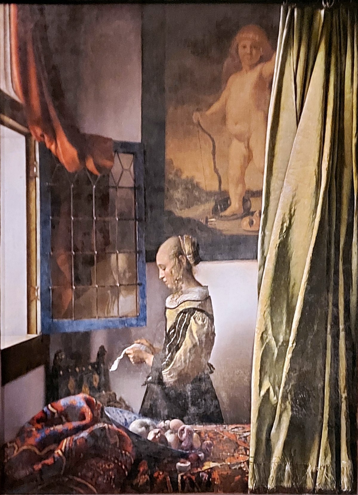 A young woman stands in profile at an open leaded window, reading a letter, her faint reflection visible in the glass. A bowl of fruit lies tipped on a rumpled carpet in the foreground, a green curtain is drawn aside at the right, and on the wall behind her hangs a large painting of Cupid holding a bow and treading on a mask.