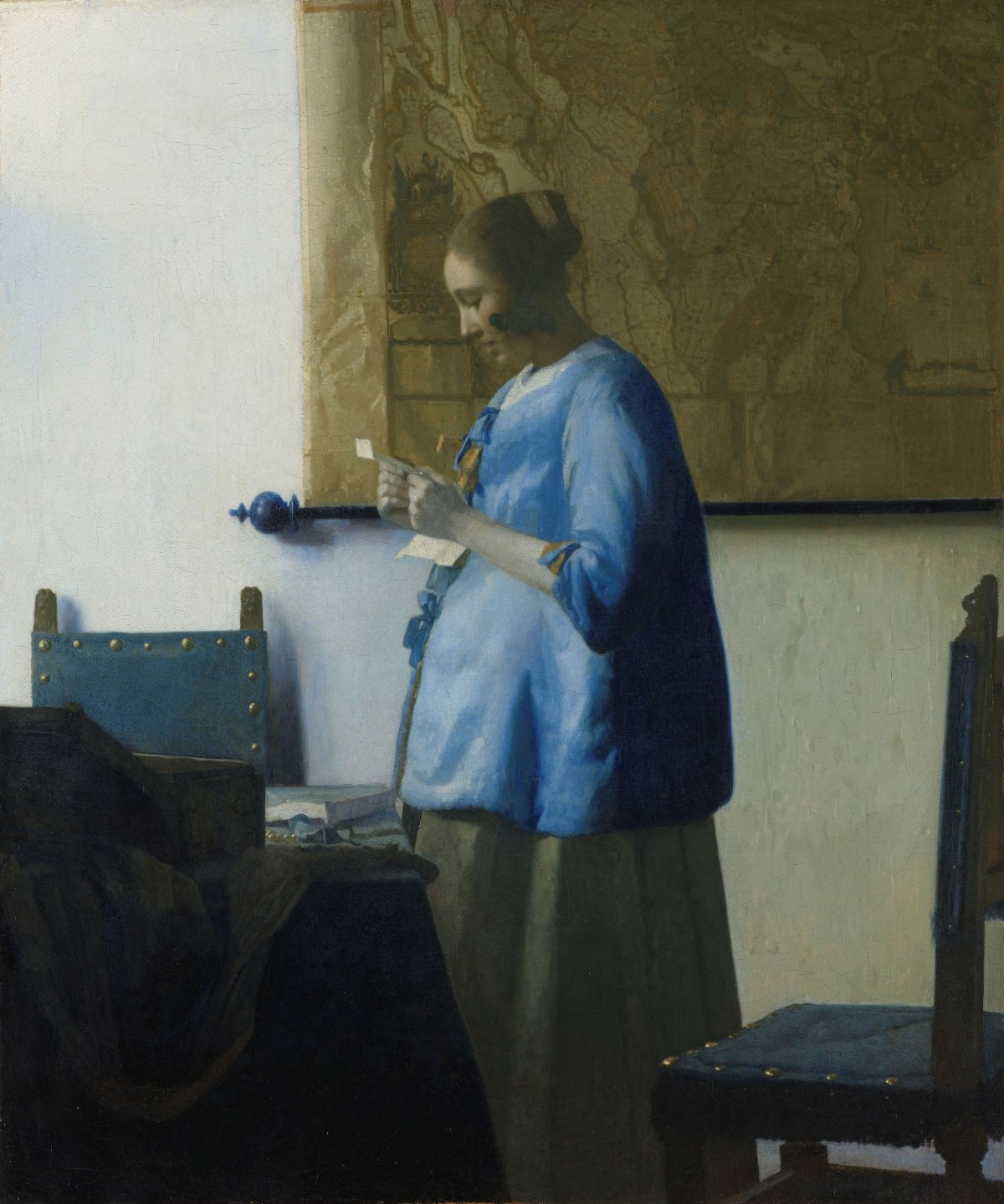 In profile, a young woman in a blue jacket reads a letter held in both hands, lit from one side. Behind her hangs a large wall map. Two chairs with brass studs stand empty in the room.