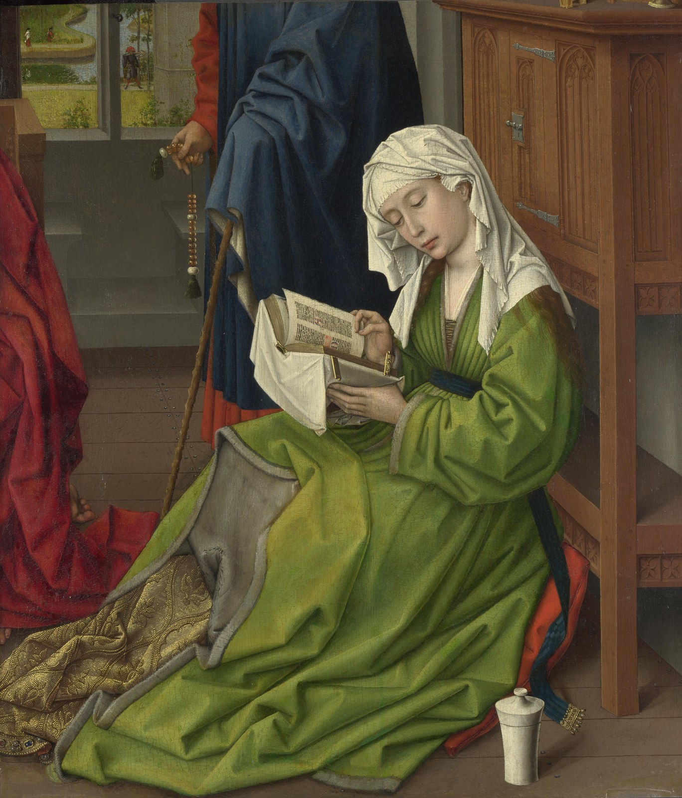 A woman in a green gown and white headdress sits on a timber floor, reading an open book held in both hands. A white ointment jar stands beside her. Behind her, a man holds a staff and a string of beads. A landscape shows through an open window.
